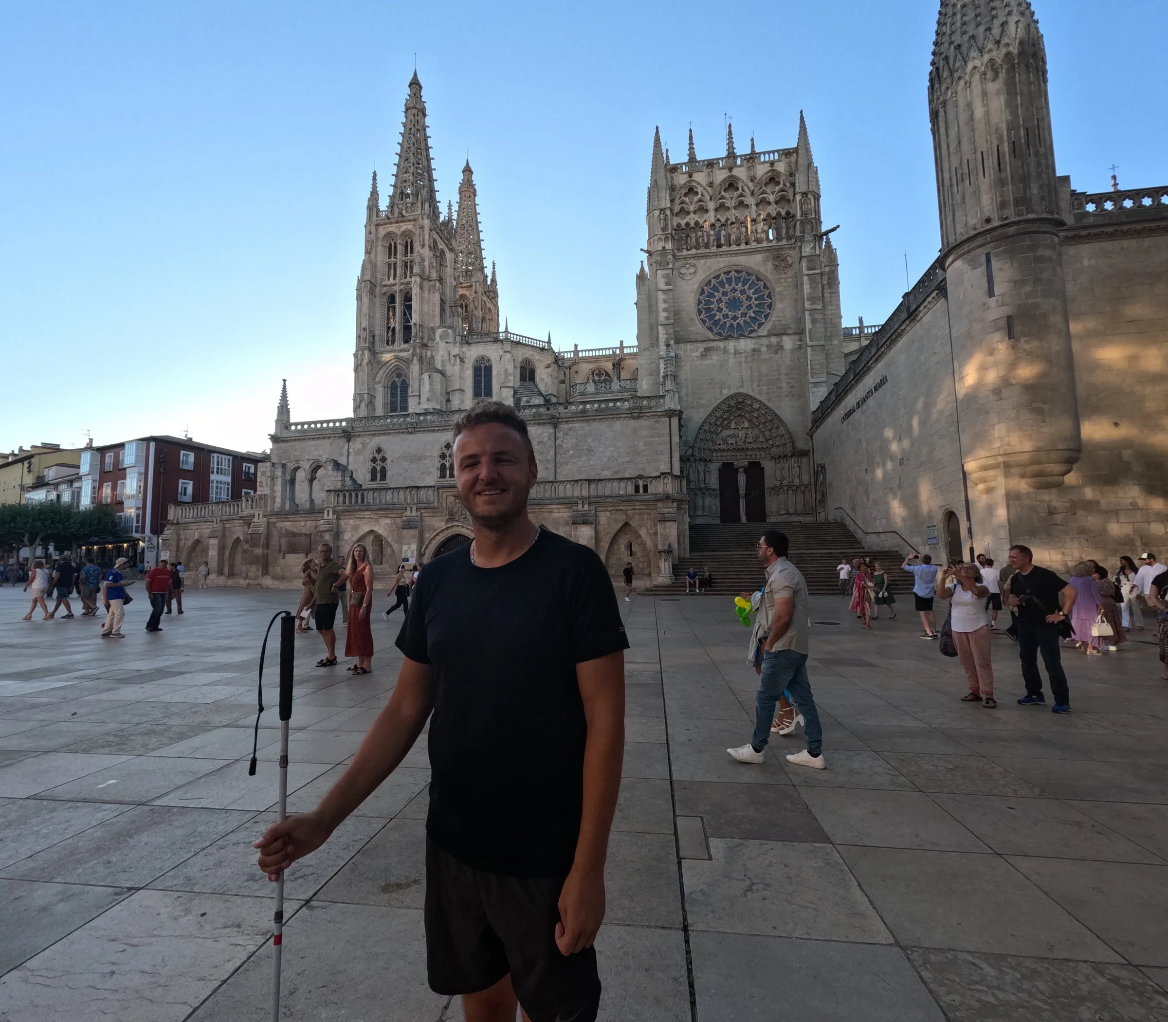 Edis smiles in front of the gothic cathedral of Burgos, wearing a black t-shirt and holding his white cane, on a sunlit square with other visitors.