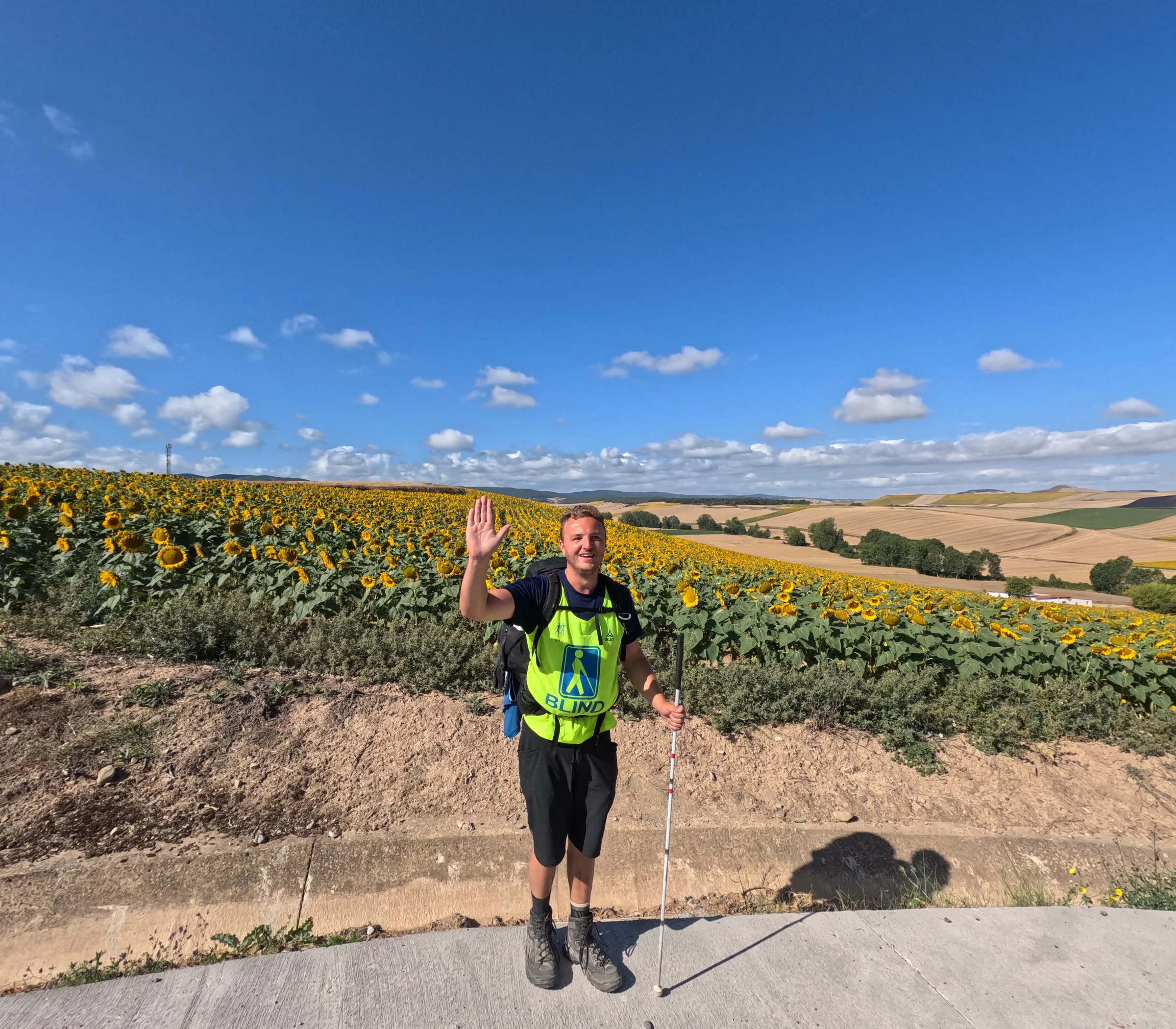 Edis smiles and waves in front of a large sunflower field under a blue sky, wearing a yellow blind-symbol vest and a backpack, cane in hand.