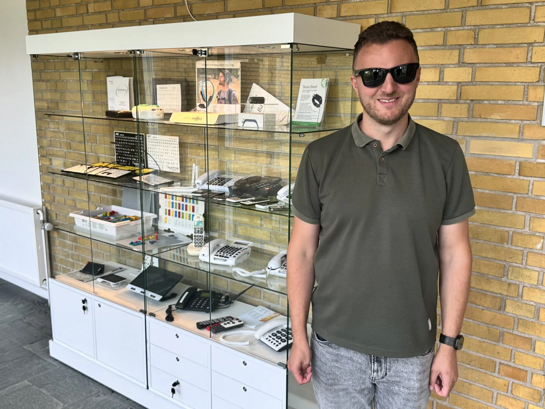 Edis in sunglasses stands smiling next to a glass display cabinet full of assistive devices: phones with big buttons, keyboards and key stickers with large letters, remote controls, bone-conduction headphones and a Sunu Band.