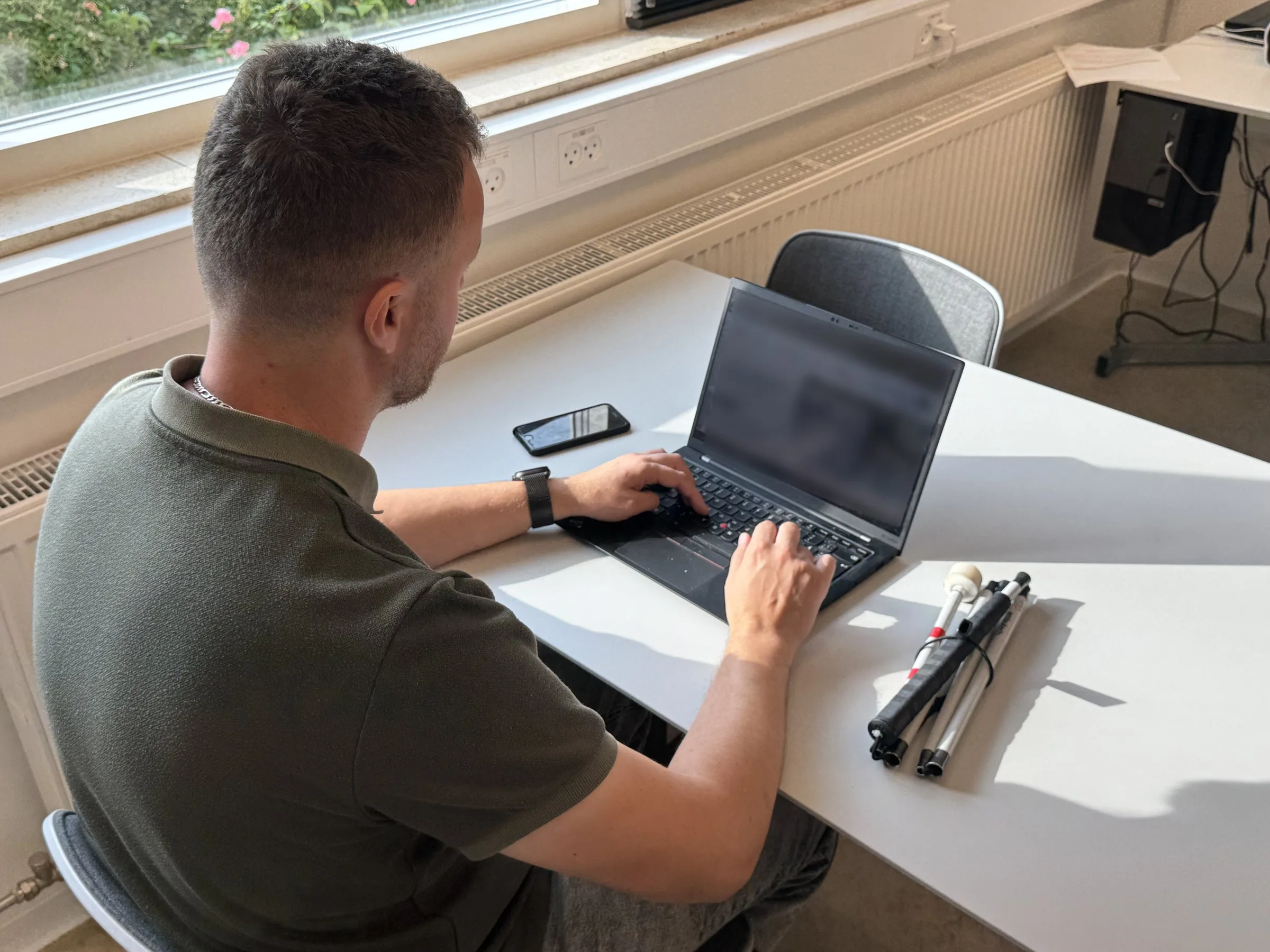 Edis seen from behind at an angle, at a white desk in bright sunlight. He types on a black laptop with both hands on the keyboard. Beside it lie his phone and a folded white cane.