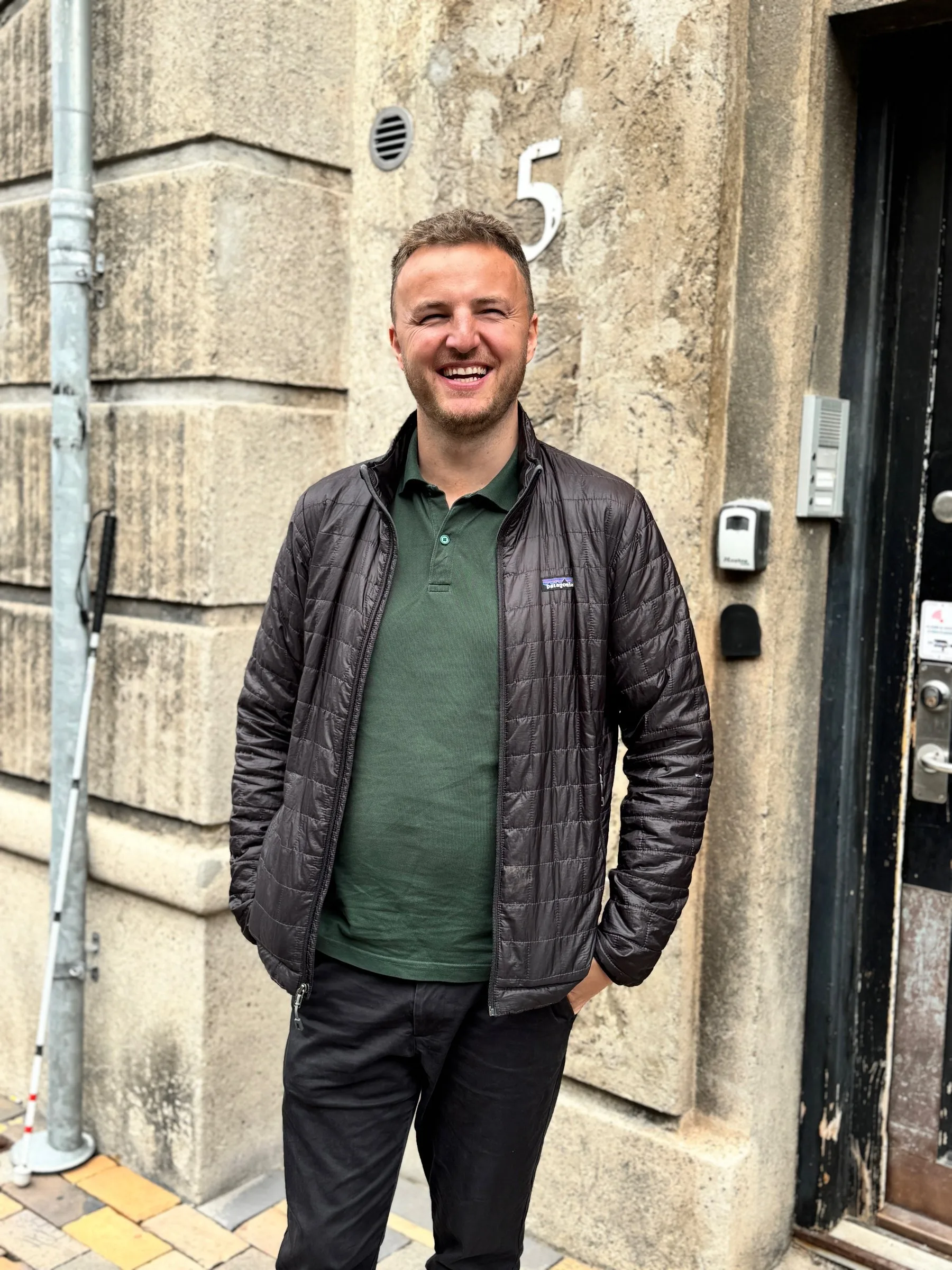 Edis laughs at the entrance to his office in Odense, wearing a dark puffer jacket and a green polo shirt. Behind him is a grey stone wall with a large white house number 5, and his white cane leans against the drainpipe on the left.