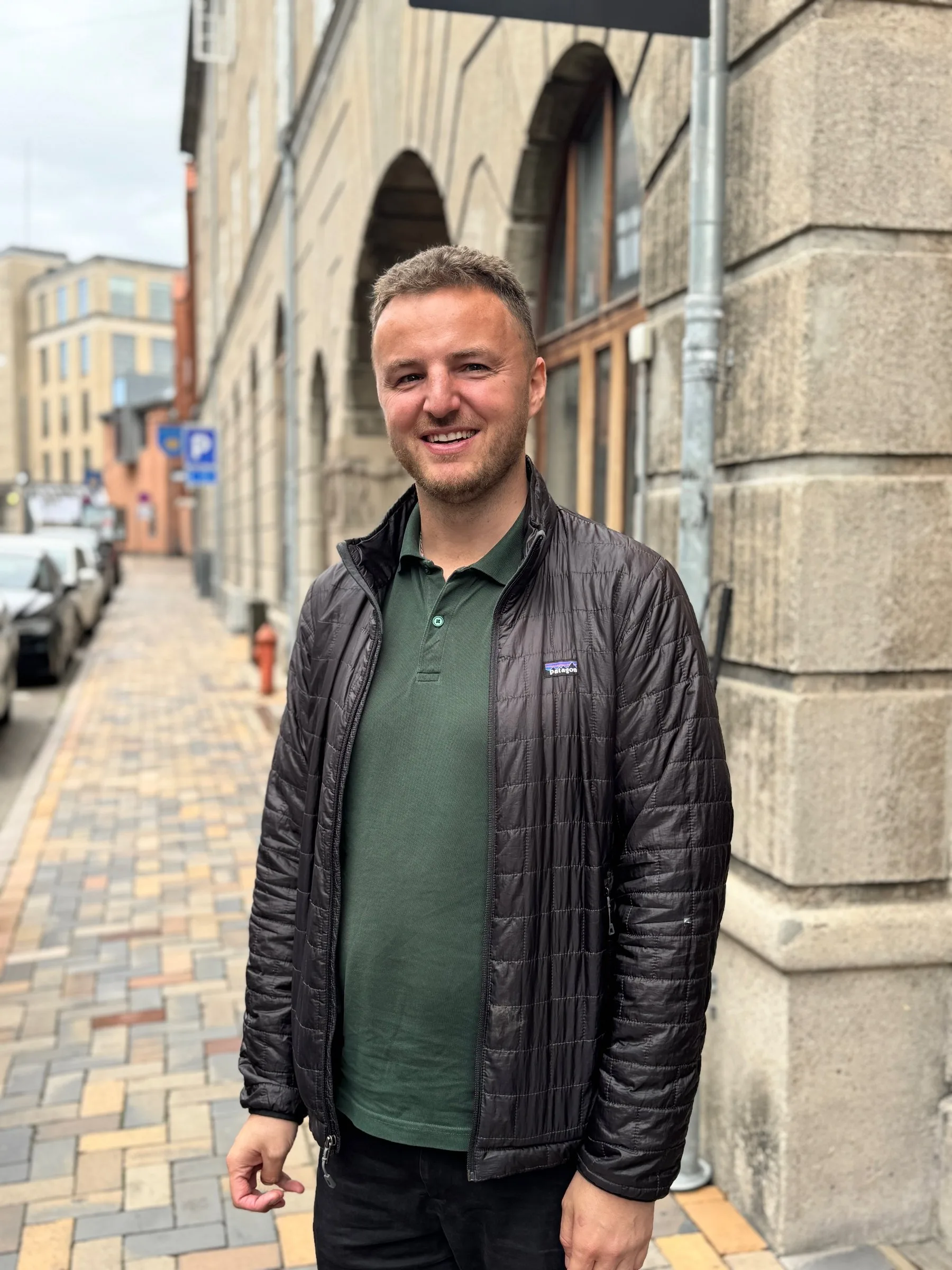 Edis står og smiler på fortovet foran sin kontorbygning på Pantheonsgade i Odense, i mørk dynejakke og grøn poloshirt. Til højre rejser bygningens grå stenfacade sig med høje buede vinduer, og bag ham løber gaden med brosten og parkerede biler videre mod flere bygninger.