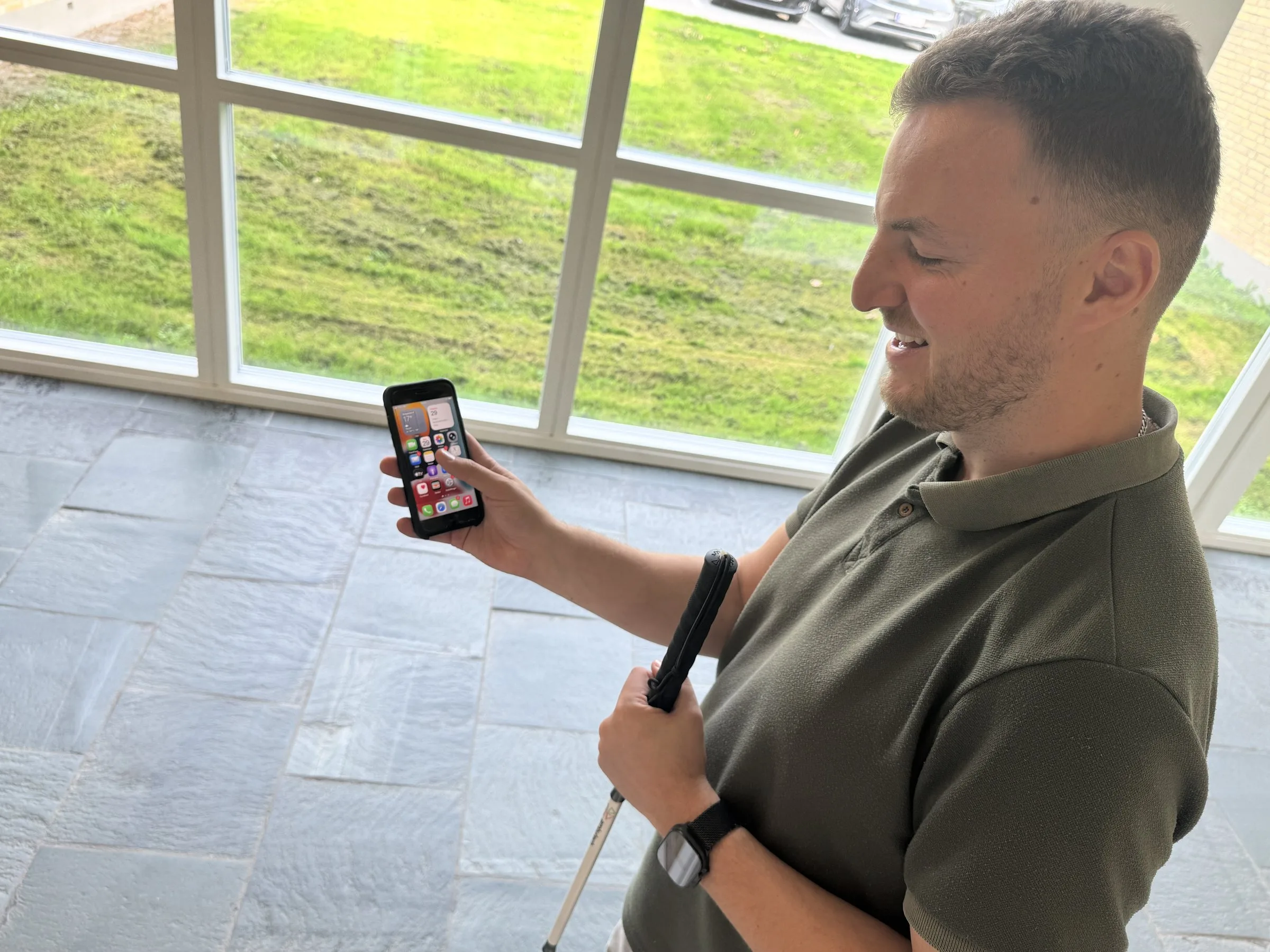 Edis stands by a large window with a lawn outside, using his iPhone. He holds the phone up in one hand, touches the screen with a finger and smiles. In his other hand he holds his white cane.