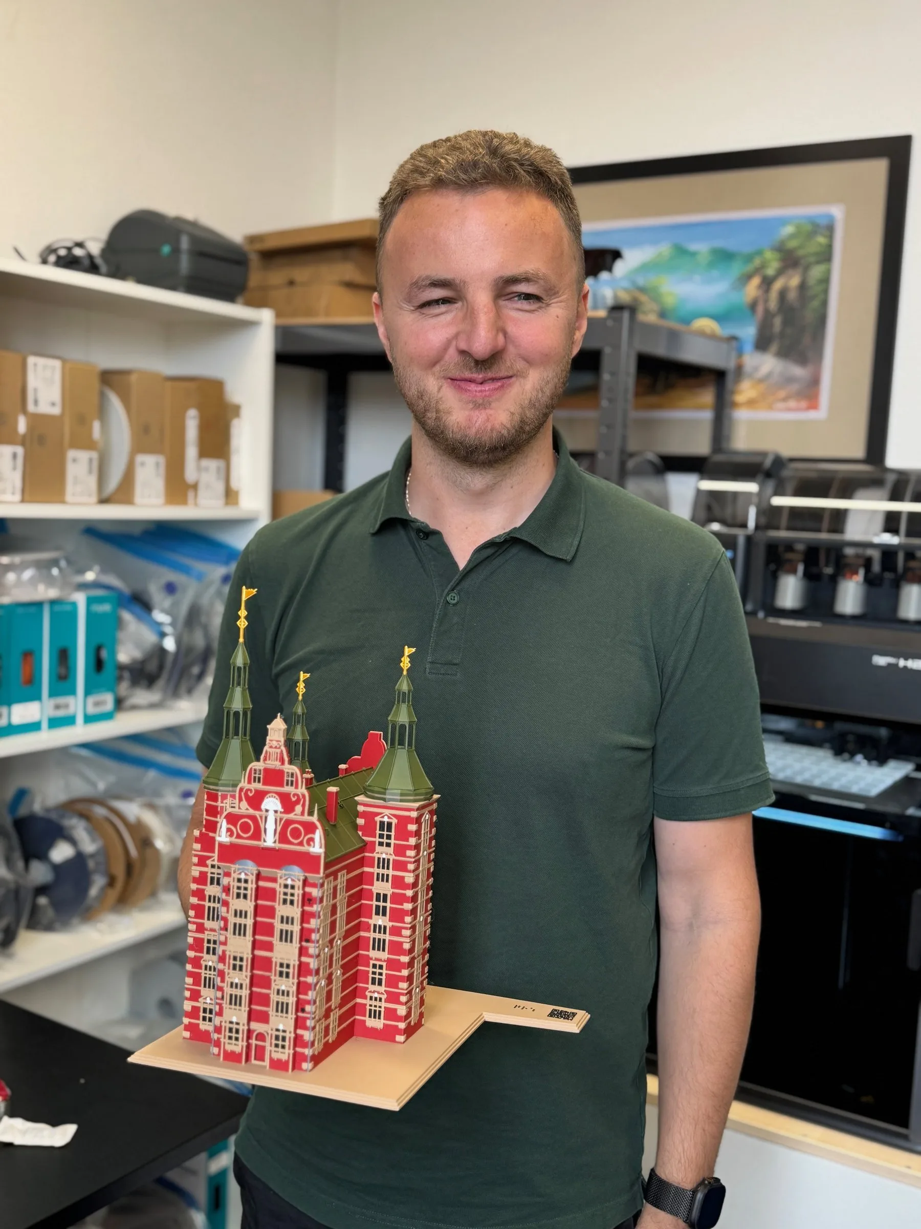 Edis smiles at the camera in his workshop, holding a coloured 3D-printed model of Rosenborg Castle with red walls and green spires. Shelves of filament and a 3D printer stand behind him.