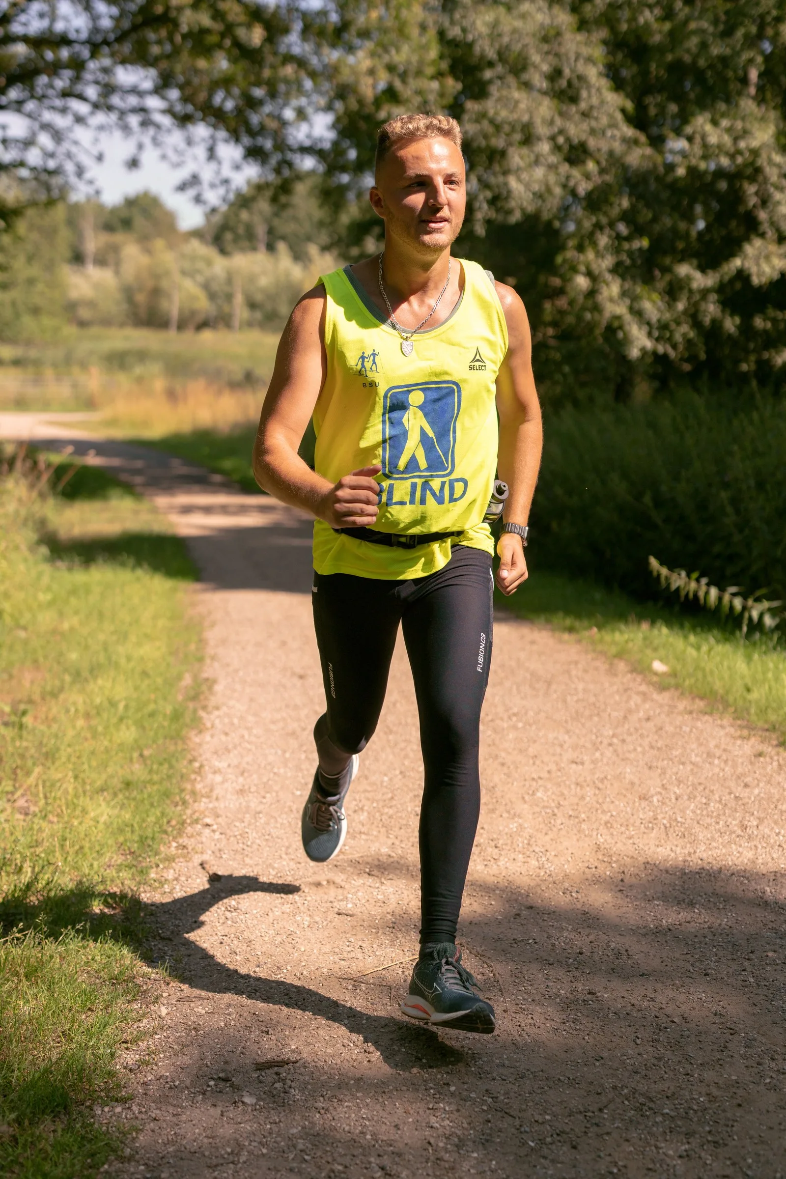 Edis runs along a sunlit path wearing a yellow blind-symbol vest and black running tights, with green bushes and trees behind him.