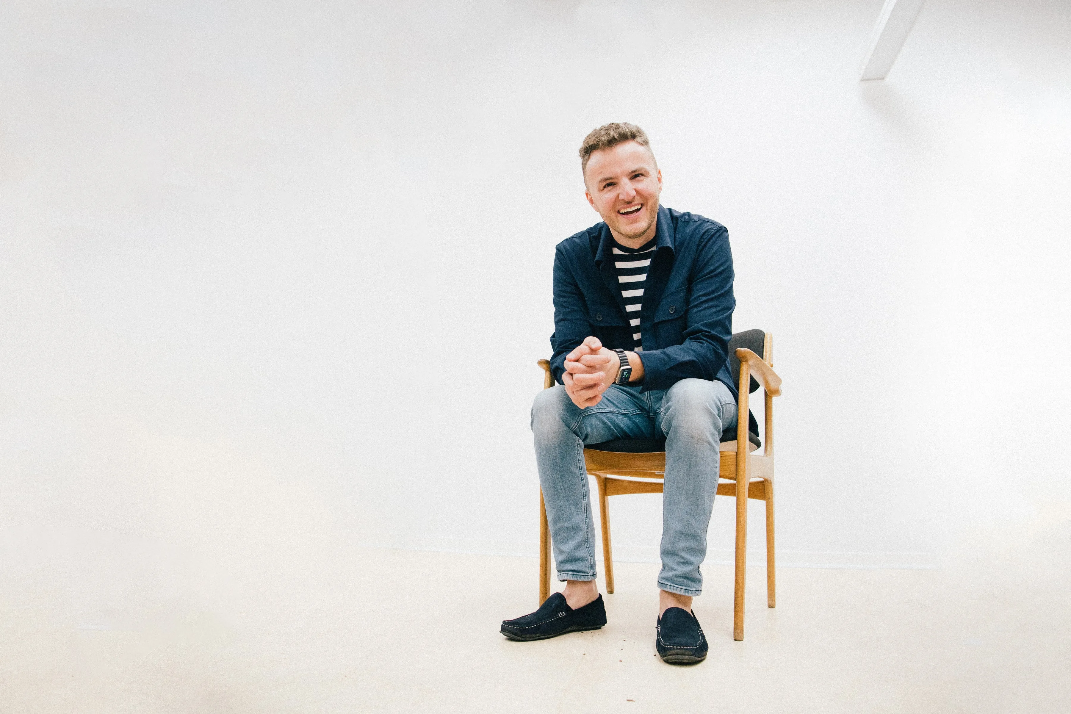 Edis sidder og smiler på en træstol i et hvidt studie, iført mørkeblå jakke, stribet trøje og lyse jeans, med hænderne foldet.