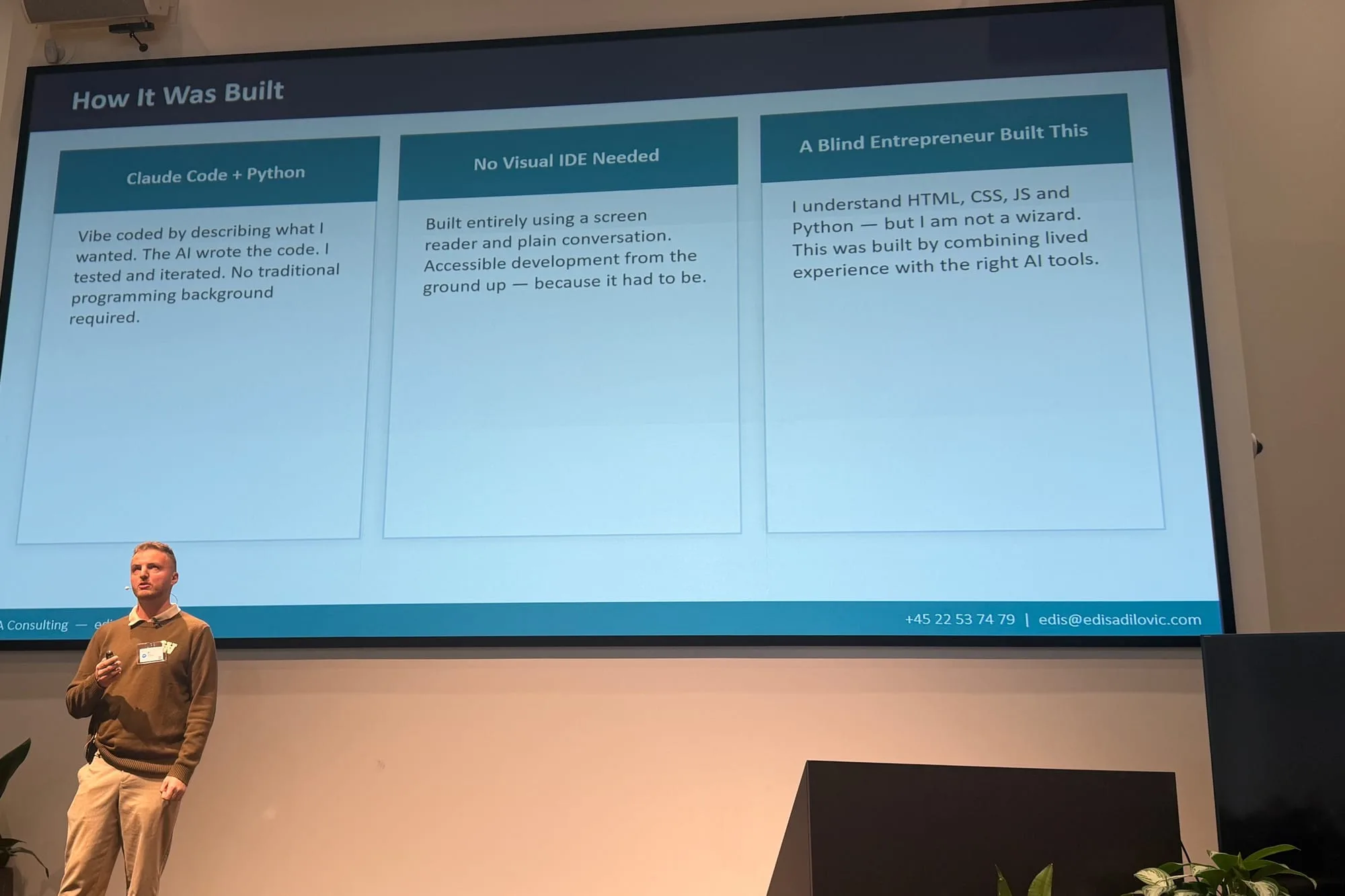 Edis står på en scene hos Microsoft med en fjernbetjening i hånden. Bag ham viser lærredet slidet Sådan blev det bygget i tre felter: Claude Code og Python, bygget ved at beskrive, hvad jeg ville have, mens AI skrev koden, og jeg testede. Intet visuelt udviklingsværktøj, kun skærmlæser og almindelig samtale. En blind iværksætter byggede det.