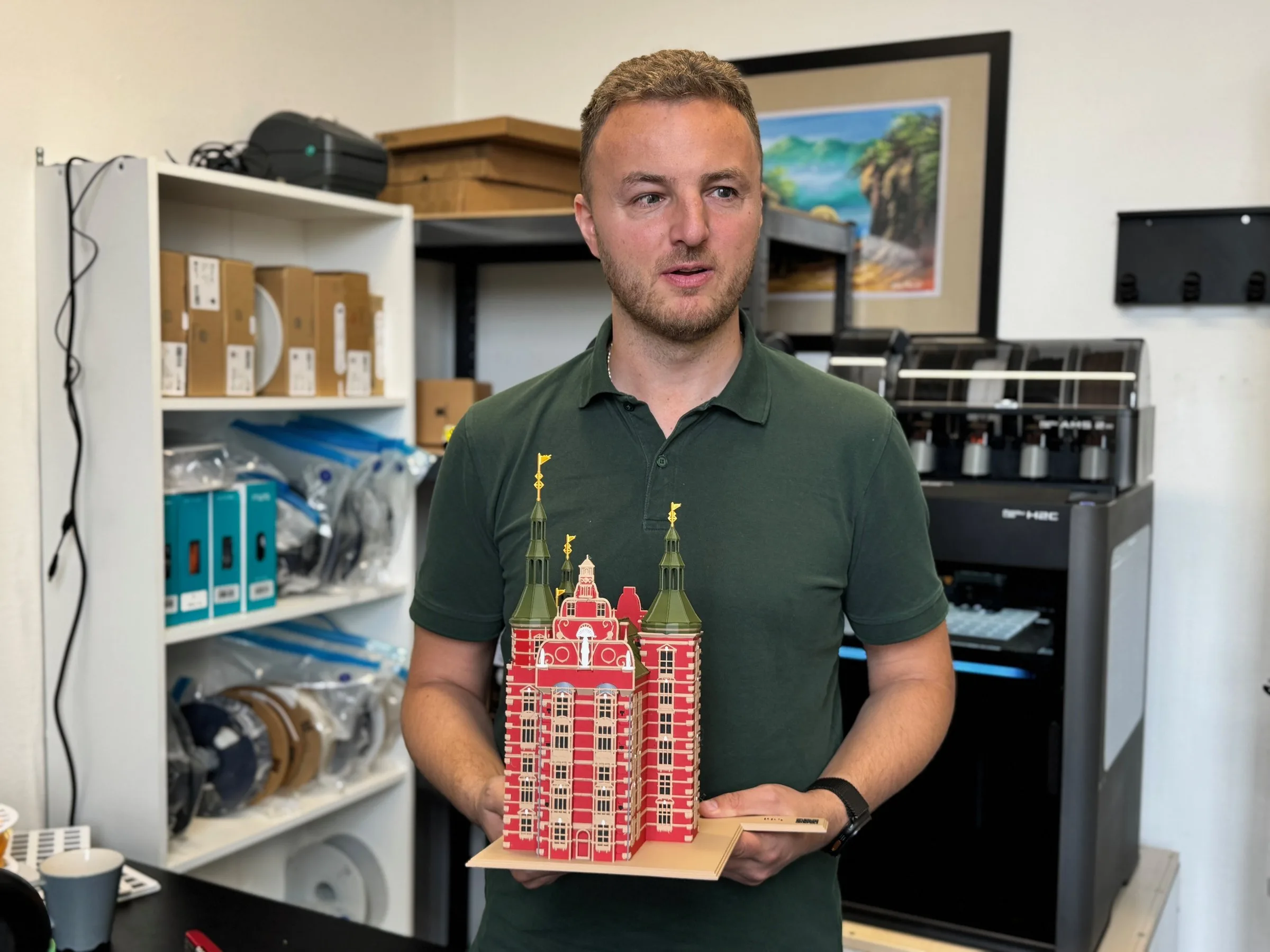 Edis stands in his workshop talking while holding a coloured 3D-printed model of Rosenborg Castle with red walls, sand-coloured windows and green spires with golden vanes. Shelves of filament spools stand behind him and a large black 3D printer on the right.