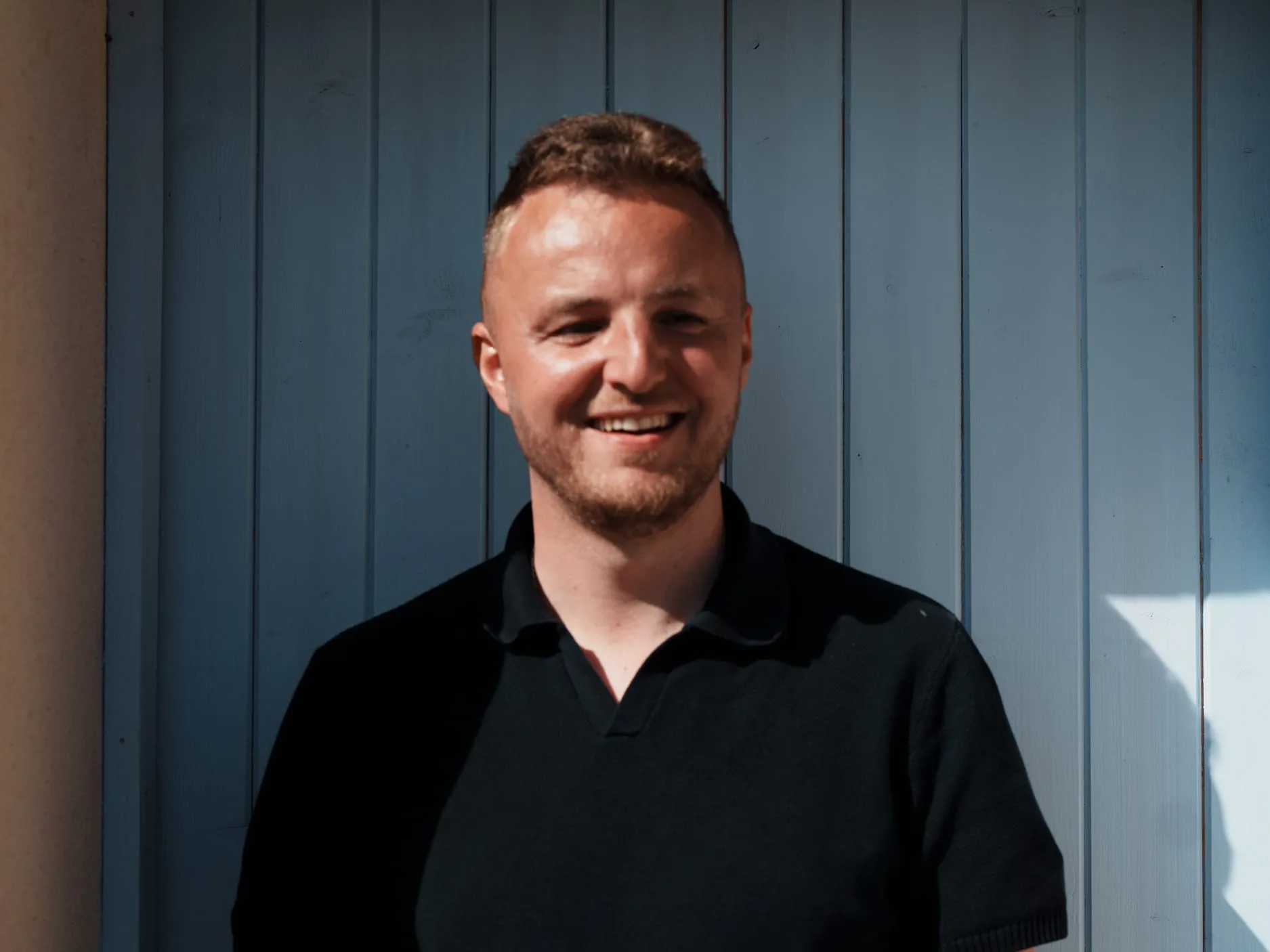 Edis smiles at the camera in front of a pale blue wooden door in bright sunlight, wearing a black polo shirt, with short light-brown hair and stubble.