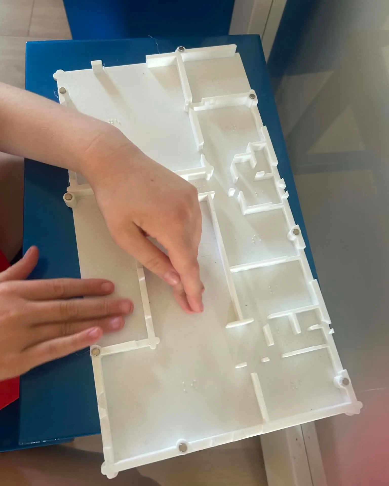 A child's hands read a white 3D-printed floor plan of a house on a blue table: raised walls, door openings and small braille marks in every room.