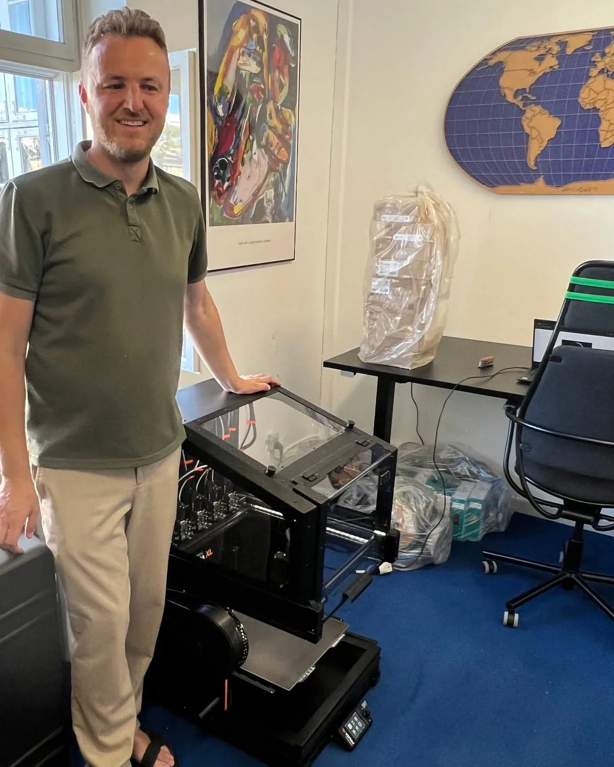 Edis stands smiling in his office with his hand resting on his black Prusa XL 3D printer. The printer enclosure has clear panels, and the print heads show behind the glass. Behind him a window, a colourful poster and a world map on the wall.
