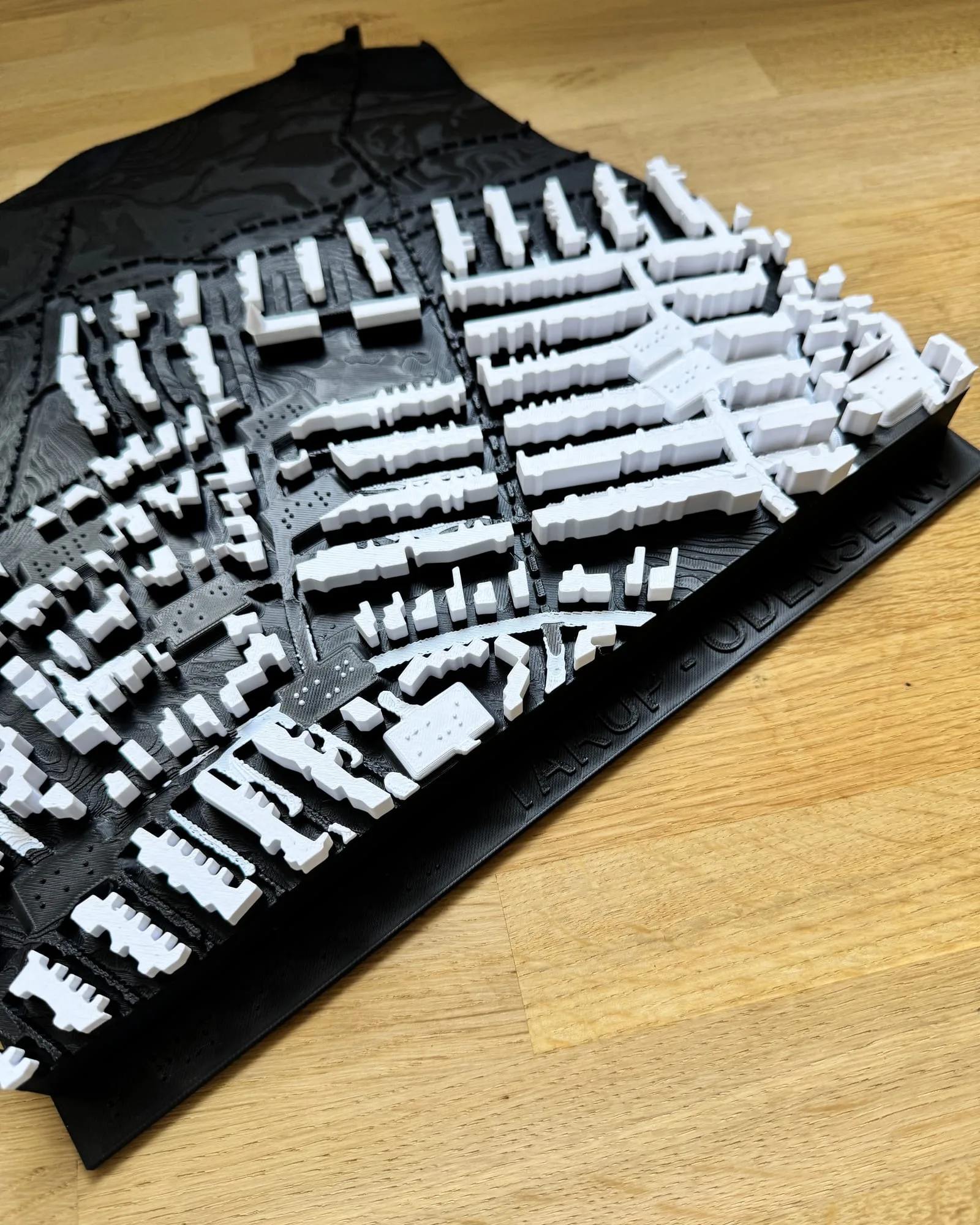 Tactile city map of Tarup in Odense NV, seen at an angle on a wooden table: white raised buildings on a black base with contour lines, streets as grooves and braille labels by the larger buildings. The edge carries the text Tarup, Odense NV.