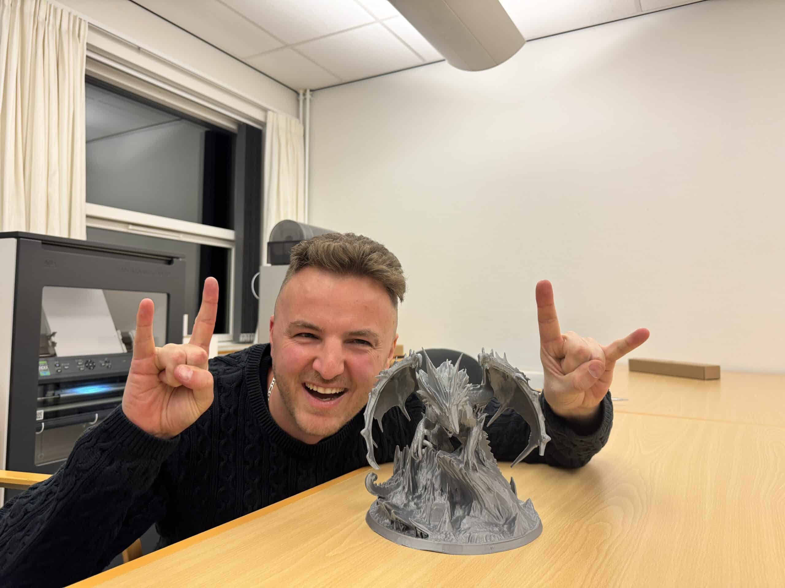 Billedet viser Edis, en ung mand med kort brunt hår og et bredt smil, siddende ved et træbord i et lyst lokale. Han har en sort kabelstrikket sweater på og laver "devil’s horn"-håndtegnet med begge hænder. Foran ham står en meget detaljeret 3D-printet model af en drage med udbredte vinger på en klippebase. I baggrunden ses en stor 3D-printer eller en lignende maskine, et vindue med hvide gardiner og nogle kontorstole. Omgivelserne ligner et værksted eller laboratorium, sandsynligvis til 3D-print eller design.