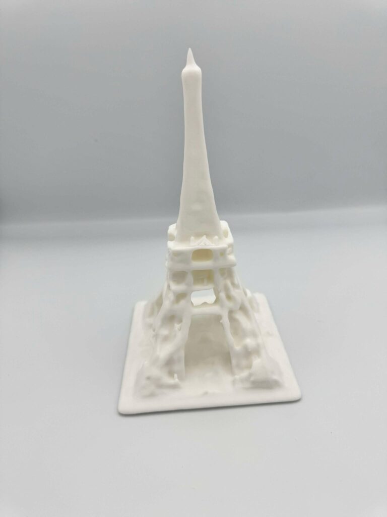Et billede af en 3D-printet model af Eiffeltårnet i hvid. Designet er stiliseret med en flydende, næsten smeltet tekstur, der giver det et abstrakt og kunstnerisk udtryk. Strukturen bevarer Eiffeltårnets ikoniske form med buer ved basen og en spids top, men de glatte og bølgende detaljer skaber en unik fortolkning af det berømte monument. Modellen er monteret på en flad base, der øger stabiliteten.