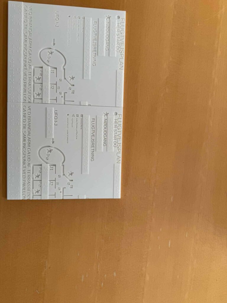 Et billede af en taktil evakueringsplan, sandsynligvis designet til brugere med synshandicap. Planen er trykt på en hvid overflade og indeholder hævet tekst, punktskrift og taktile symboler. Vigtig information som nødudgange, flugtveje og samlingspunkter er tydeligt markeret med både taktile diagrammer og prægede etiketter. Layoutet inkluderer løbende figurer og pile, der angiver retninger. Kombinationen af punktskrift og visuelle elementer sikrer tilgængelighed for både seende og blinde. Planen ser ud til at være monteret eller udstillet på en træoverflade.
