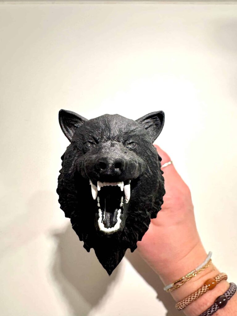 Et nærbillede af en sort ulveformet knivknop med detaljeret pelstekstur og en åben mund med markante hvide hugtænder. Håndtaget holdes af en hånd iført flere armbånd mod en hvid baggrund.