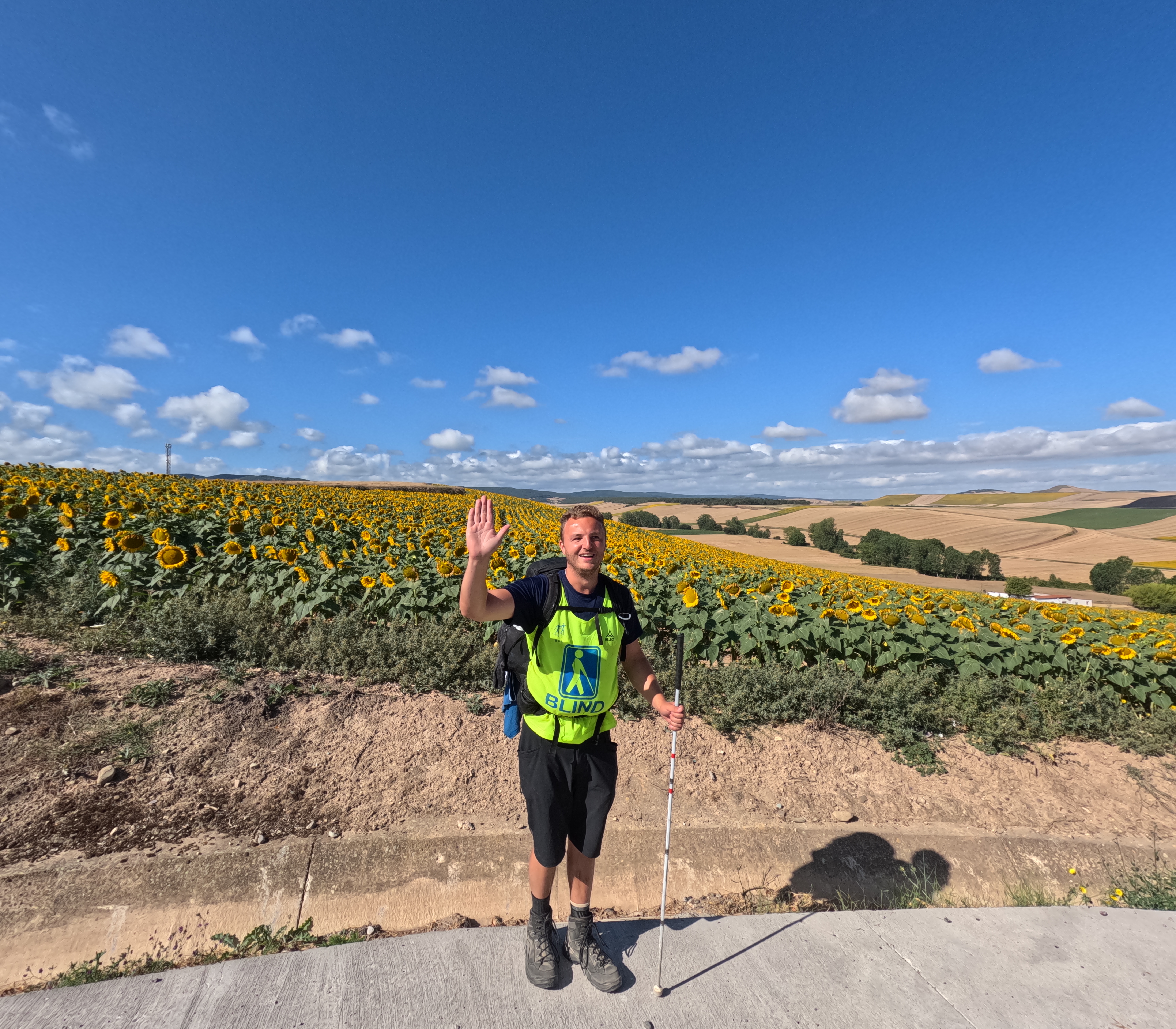 Edis smiler og vinker foran et stort solsikkefelt iført gul blindevest, rygsæk og stok under en blå himmel.